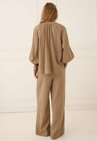 A woman viewed from the back wearing a matching beige and brown checkered two-piece set consisting of a long-sleeve blouse with balloon sleeves and wide-leg trousers, set against a clean white paneled studio background.
