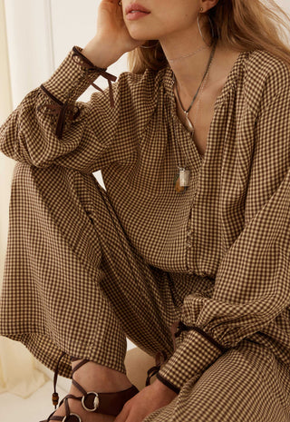 A woman wearing a brown and beige gingham long-sleeve jumpsuit with tie details on the cuffs, paired with brown strappy sandals and silver jewelry.